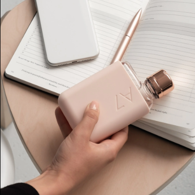 Frasco de perfume rosa com tampa dourada na mão, telemóvel branco e caderno aberto sobre mesa de madeira