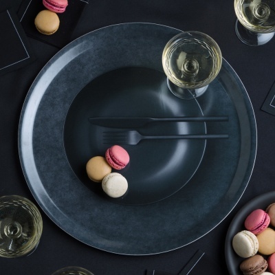 Conjunto de loiça escura com talheres pretos, macarons coloridos e copos de bebida sobre mesa preta