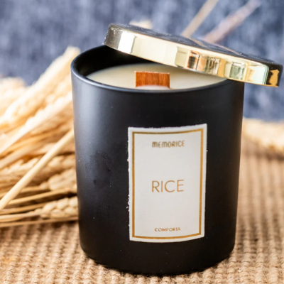 Vela perfumada preta com tampa dourada e espigas de trigo ao fundo