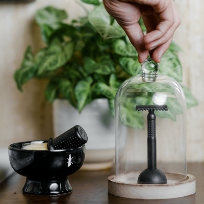 Relógio de mesa com suporte preto e tampa de vidro, mão levantando a tampa, pilão preto e planta verde