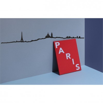 Cartaz vermelho com a palavra PARIS encostado a parede cinzenta com linha do horizonte de Paris