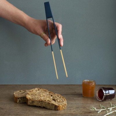 Pinça azul e amarela segurada à mão sobre mesa com pão e frascos de compota
