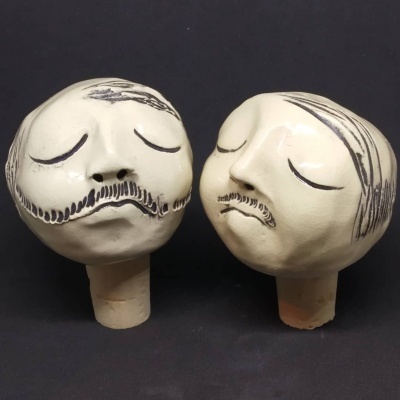 Cabeças de cerâmica com rosto masculino e olhos fechados, bigodes e cabelo desenhados, sobre base cilíndrica.