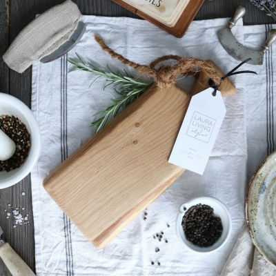 Tábua de madeira clara com etiqueta e ervas aromáticas sobre pano branco e utensílios de cozinha