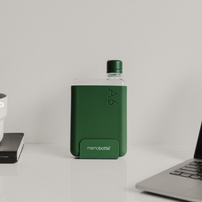 Garrafa de água plana verde A6 memobottle em superfície branca entre livro e portátil