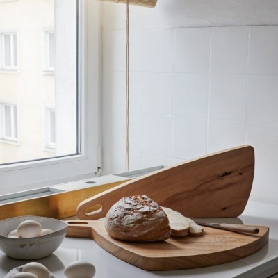 Tábuas de cortar em madeira com pão fatiado, ovos em tigela e toalha cinzenta numa bancada branca junto a janela com persiana.