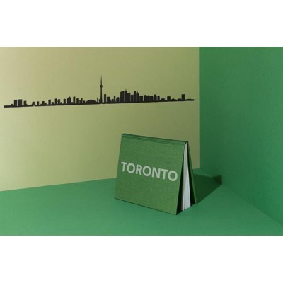 Livro verde de capa dura com 'TORONTO' em branco sobre superfície e parede verdes, com silhueta preta da linha do horizonte de Toronto na parede creme