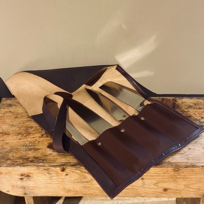 Conjunto de facas em bolsa de couro castanho escuro sobre mesa de madeira.