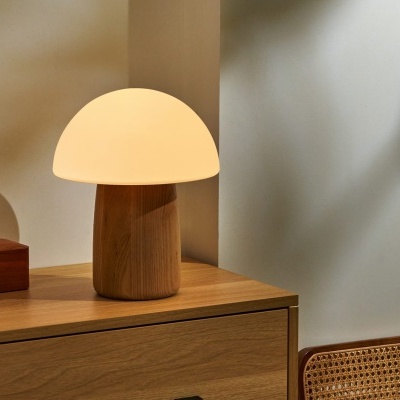 Candeeiro de mesa em madeira clara com cúpula branca sobre móvel de madeira clara e cadeira com encosto em rattan