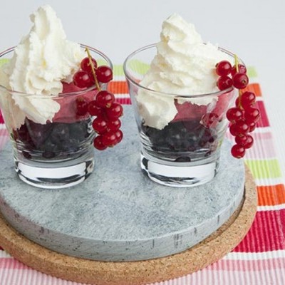 Dois copos com sobremesa de creme branco e frutas vermelhas sobre base cinza e suporte de cortiça numa toalha de mesa colorida.