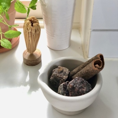 almofariz branco com pedras e pau de canela, suporte branco com utensílios de madeira, escova de chá de bambu, planta verde em vaso de barro
