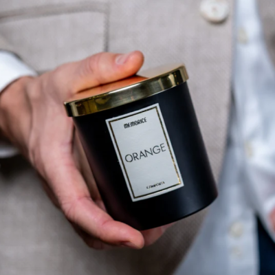 Vela aromática preta com etiqueta 'THE AROMANCE ORANGE' e tampa dourada nas mãos de uma pessoa