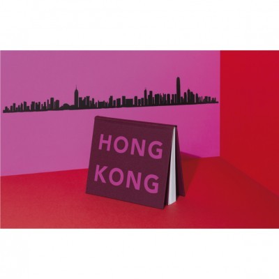 Livro com capa preta e letras cor-de-rosa HONG KONG sobre superfície vermelha e parede cor-de-rosa com autocolante preto de skyline