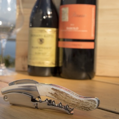 Saca-rolhas com cabo de osso sobre mesa de madeira, garrafas de vinho e taças ao fundo
