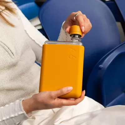 Garrafa de vidro com capa silicone laranja e tampa amarela na mão de mulher, cadeira azul ao fundo