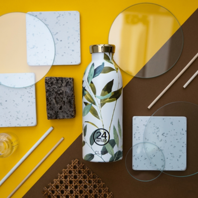 Garrafa térmica decorada com padrão de folhas verdes e tampa dourada, rodeada por materiais diversos sobre fundo amarelo e castanho.