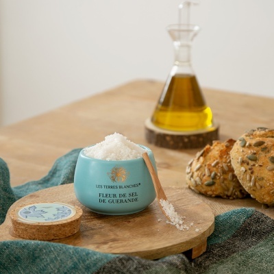 Tigela azul com flor de sal, pauzinho de madeira, pães com sementes, tábua de madeira e garrafa de azeite