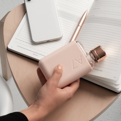 frasco de perfume rosa claro com tampa dourada em mão num ambiente com caderno e telemóvel