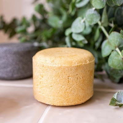 Champô sólido amarelo com textura granulada e plantas verdes no fundo