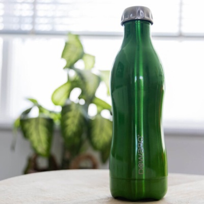 Garrafa metálica verde com tampa prateada numa mesa com plantas e janela ao fundo