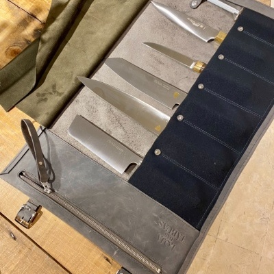 Conjunto de facas de cozinha em estojo de tecido azul e bolso de couro cinzento, sobre mesa de madeira
