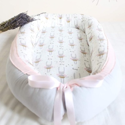 Ninho de bebé branco e rosa com padrão de coelhos e laço rosa, com ramo de lavanda ao lado