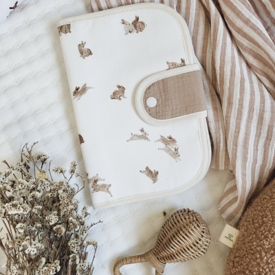 Estojo branco com coelhinhos castanhos e fecho bege,num ambiente com tecido listrado, flores secas e chocalho de vime.