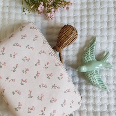 Carteira rosa com padrão floral, objeto de vime, pássaro cerâmico verde e flores sobre tecido branco.