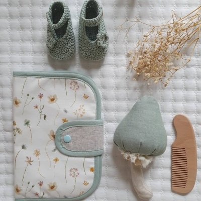 Acessórios para bebé, incluindo sapatinhos de crochet verdes, carteira floral, brinquedo de cogumelo em peluche e pente de madeira sobre manta branca.