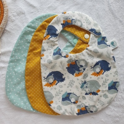 Três babetes para bebés com padrões coloridos e fechos de pressão.