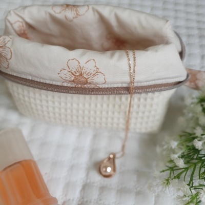 Cesto com colar dourado, frasco translúcido e flores brancas sobre tecido branco texturizado