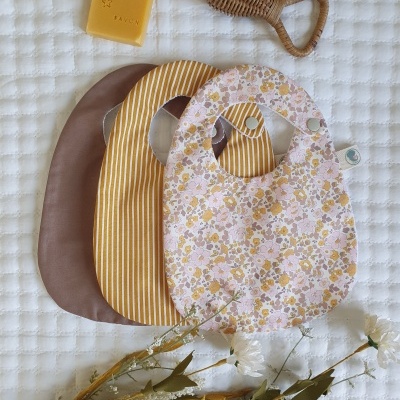 Três babetes de tecido com padrões diferentes, sabonete amarelo, escova de cabelo de madeira e flores secas sobre tecido branco acolchoado