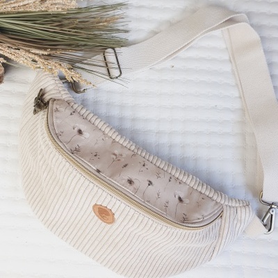 Bolsa pochete creme com padrão floral e alça ajustável sobre fundo branco texturizado com planta seca