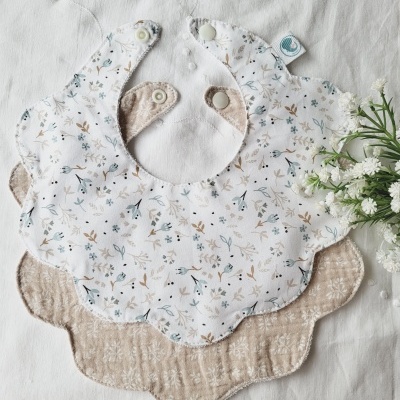 Dois babetes para bebé em tecido com padrão floral e bege, em forma de pétalas, sobre fundo branco com flores ao lado.