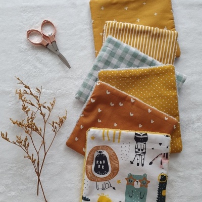 Seis panos decorativos com padrões variados, tesoura rosa e ramo seco sobre toalha branca