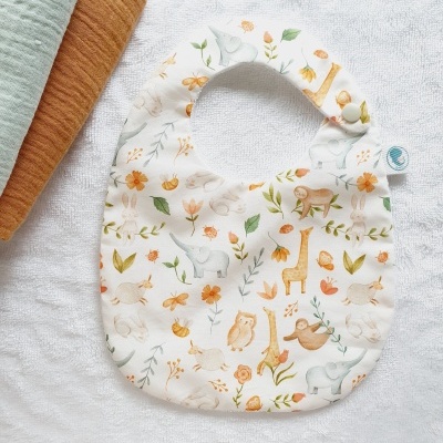 Babete para bebé com padrão de animais e flores numa superfície branca texturizada com toalhas coloridas ao lado