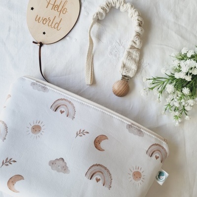 Bolsa de pano branca com padrões de sol, lua, nuvem e arco-íris em castanho claro junto a um balão de madeira com texto 'Hello world' e flores brancas