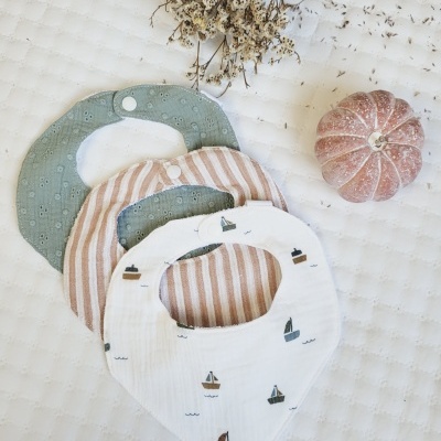 Três babetes de bebé em tecido com padrões diversos, flores secas e abóbora pequena sobre fundo branco acolchoado.