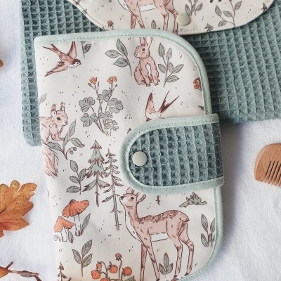 Carteira ou estojo de tecido com padrão de animais da floresta e fecho verde sobre tecido branco e folhas secas