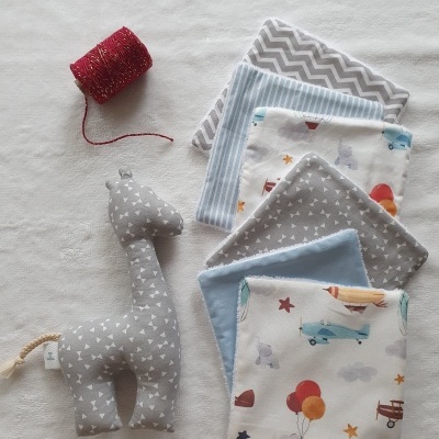 Brinquedo giraffe de peluche cinzento e panos infantis com estampados variados sobre fundo branco.