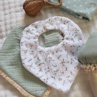 Babete branco com padrão floral, paninhos verdes, chocalho de vime, sobre colchão branco acolchoado