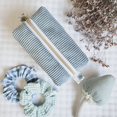 Estojo cinzento claro com fecho dourado, dois scrunchies azuis, ramo de flores secas e brinquedo em forma de cogumelo em tecido.