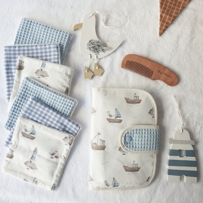 Conjunto de panos de tecido azul e branco com estampas náuticas, escova de cabelo em madeira, porta-fraldas e enfeites de gaivota e casa de praia sobre toalha branca