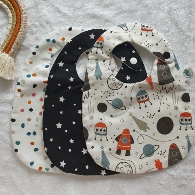 Três babetes de tecido com padrões variados, dispostos sobre um tecido cinza claro.