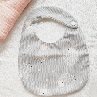 Babete cinzento com padrão de ovelhas, fecho de pressão branco, sobre toalha branca com tecido rosa ao lado