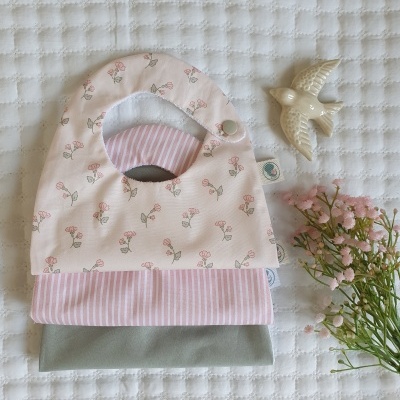 Três babetes para bebé com padrões florais, riscas e liso, sobre tecido branco acolchoado, com flores e uma ave decorativa