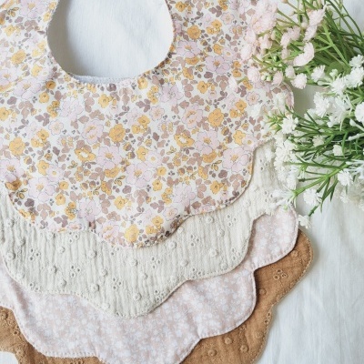 Babetes com padrões florais e bordado inglês, em tons suaves, dispostos em leque sobre tecido branco com flores ao lado