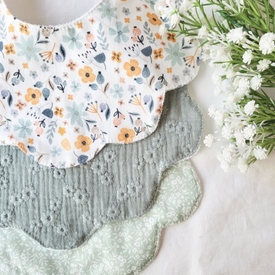 Três babetes em tecido com padrão floral e bordas onduladas, e flores brancas ao lado.