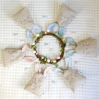 Seis sacos de coelho em tecido bege com orelhas à risca e expressões bordadas em círculo à volta de uma coroa decorativa com ovos pastel sobre colcha branca acolchoada