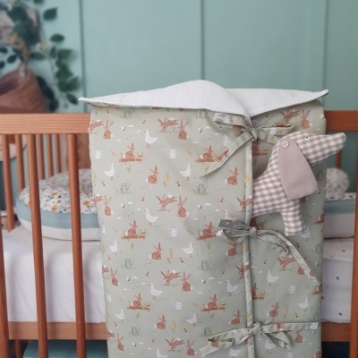 Capa almofadada bege com estampado de animais presa a berço de madeira clara com adereço coelho xadrez